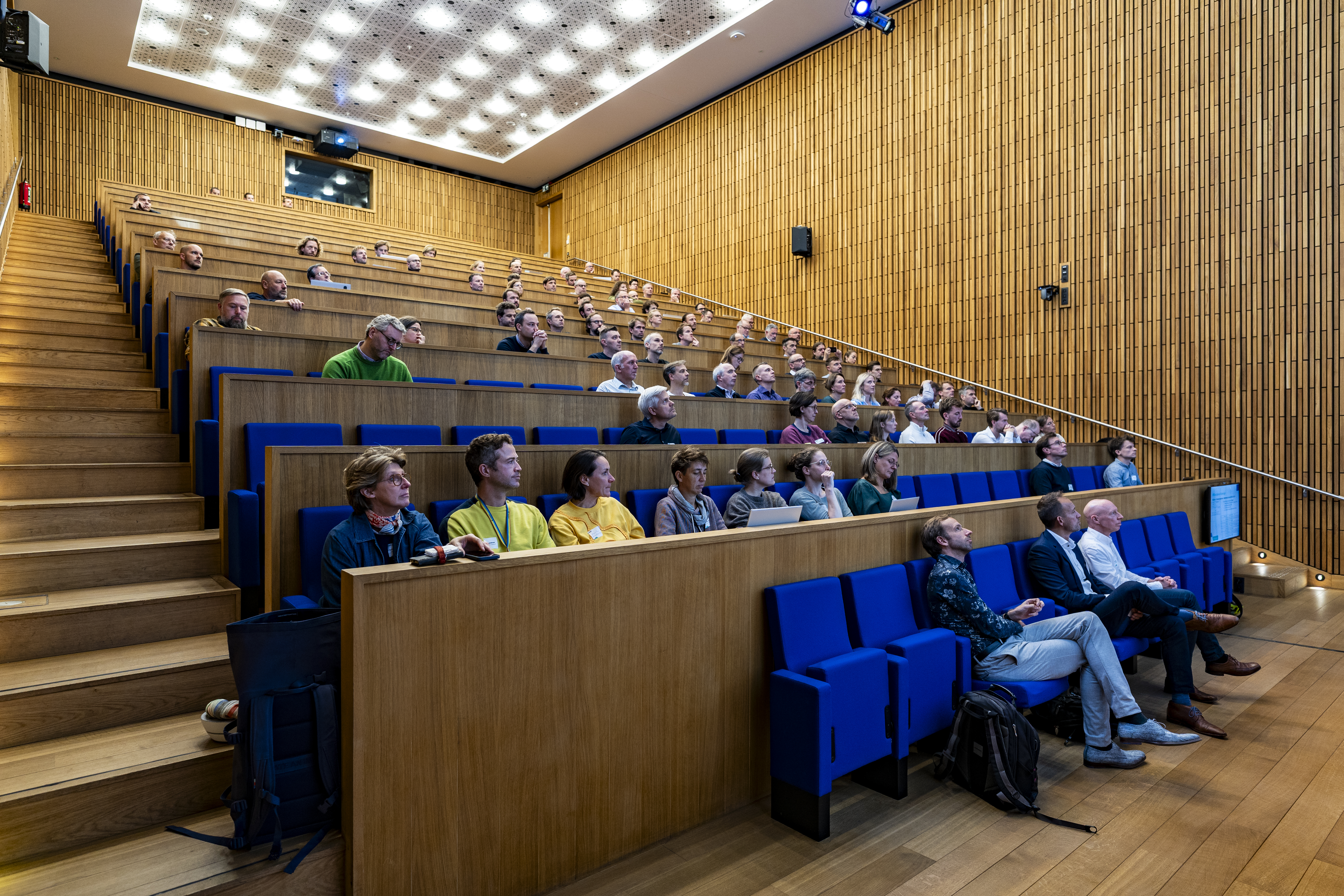 auditorium met houten muurbekleding, blauwe stoelen waarop mensen zitten